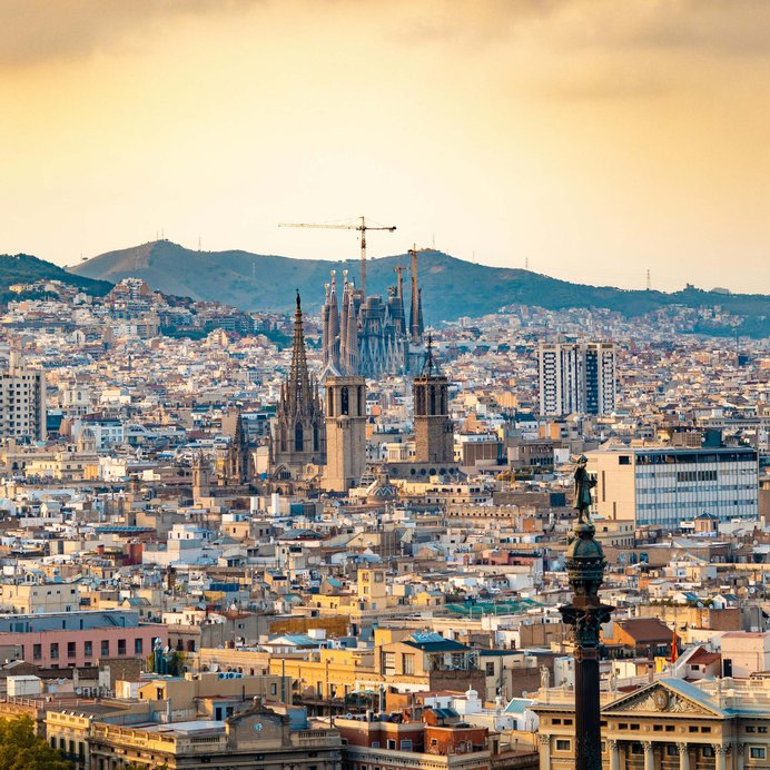 Barcelona skyline at sunset, with the spires of Barcelona Cathedral and the Sagrada Família rising above dense rooftops, hills behind.