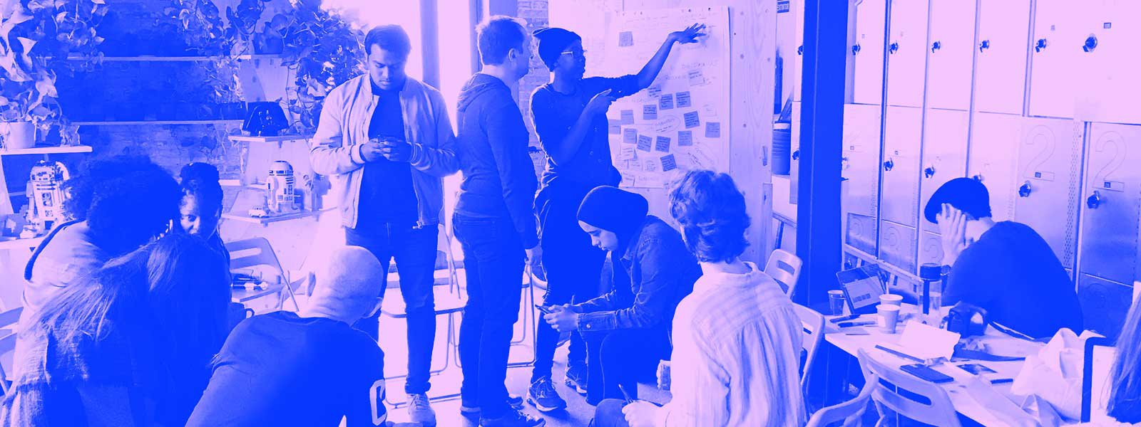 A group of several people, some seated and some standing, planning and discussing MozFest together in a sunlight room with large wooden shelves full of plants, metal storage lockers, a white board, and sticky notes on the wall.